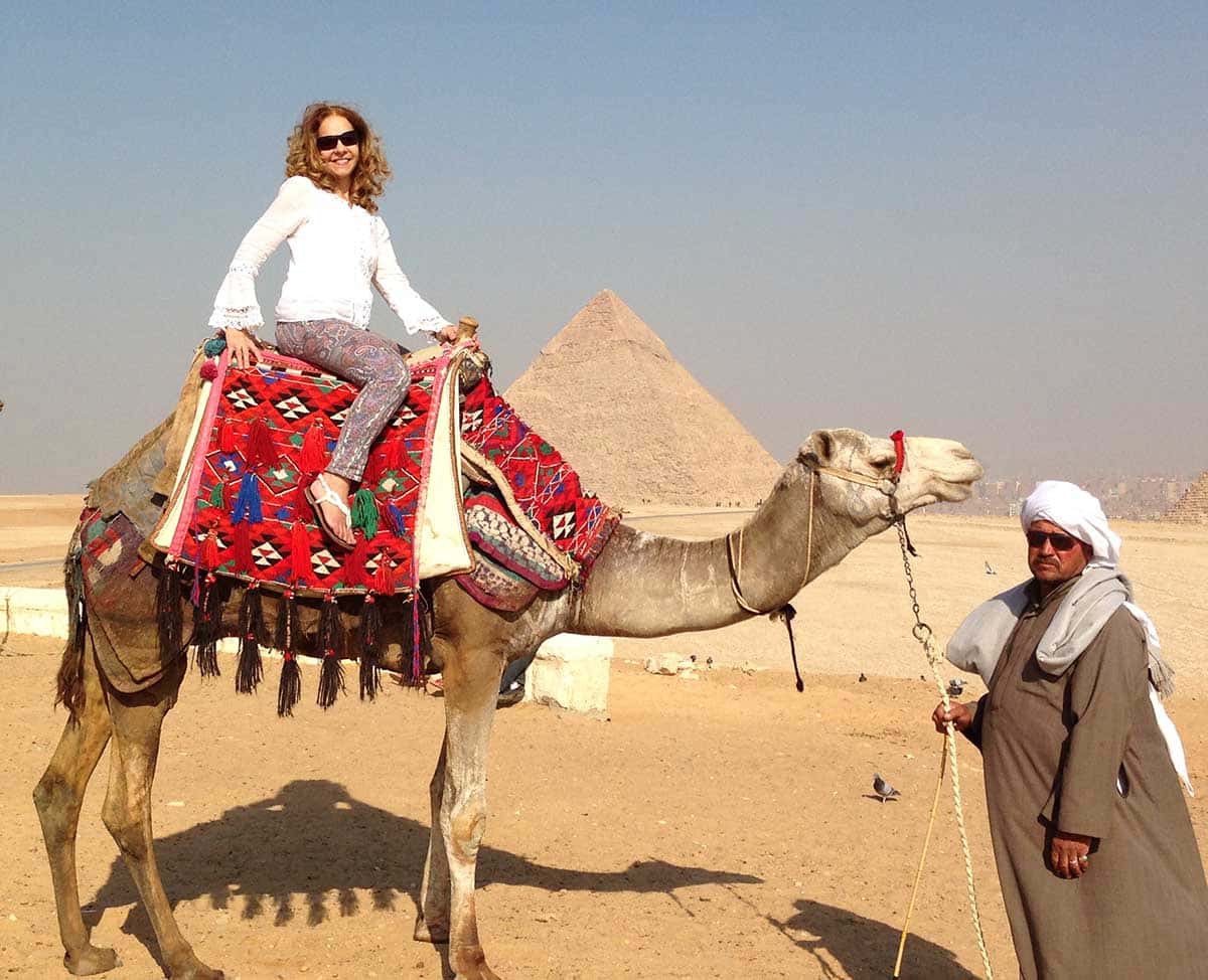 Moça loira montada num camelo e um arabe segurando o camelo. As piramides do egito estão ao fundo.