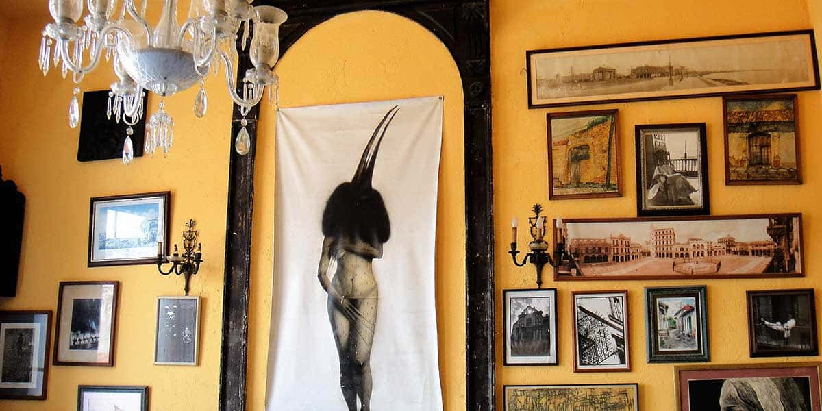 Foto de uma Parede de cor amarela cheia de quadros, com um lustre em cristal do restaurante Paladar La Guarida, dicas de onde beber e comer em Havana