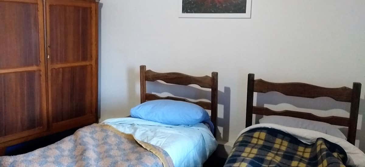quarto mostrando 2 camas, já com cobertores e travesseiros um armário de madeira encostado na parede e um quadro dependurado do Santuário do Caraça, Santuário do Caraça: programa de final de semana