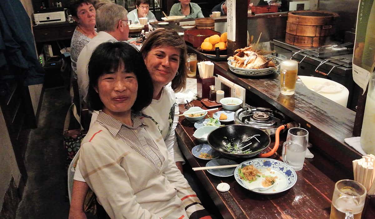 Duas moças, uma japonesa de nome Miki kuwano e outra estrangeira de Patricia Lamounier estão sentadas em um balcao de restaurante, cheio de pratos de comida a frente delas, e pessoas em volta do balcao, na região de Pont Cho , kyoto, no balcão em Pont Cho