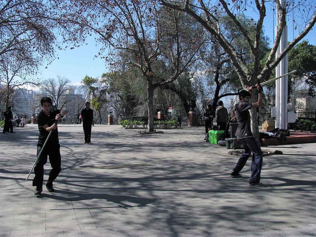 Rapazes vestidos de preto fazem exercícios marciais em parque com árvores, bancos e mais pessoas: o que fazer em santiago