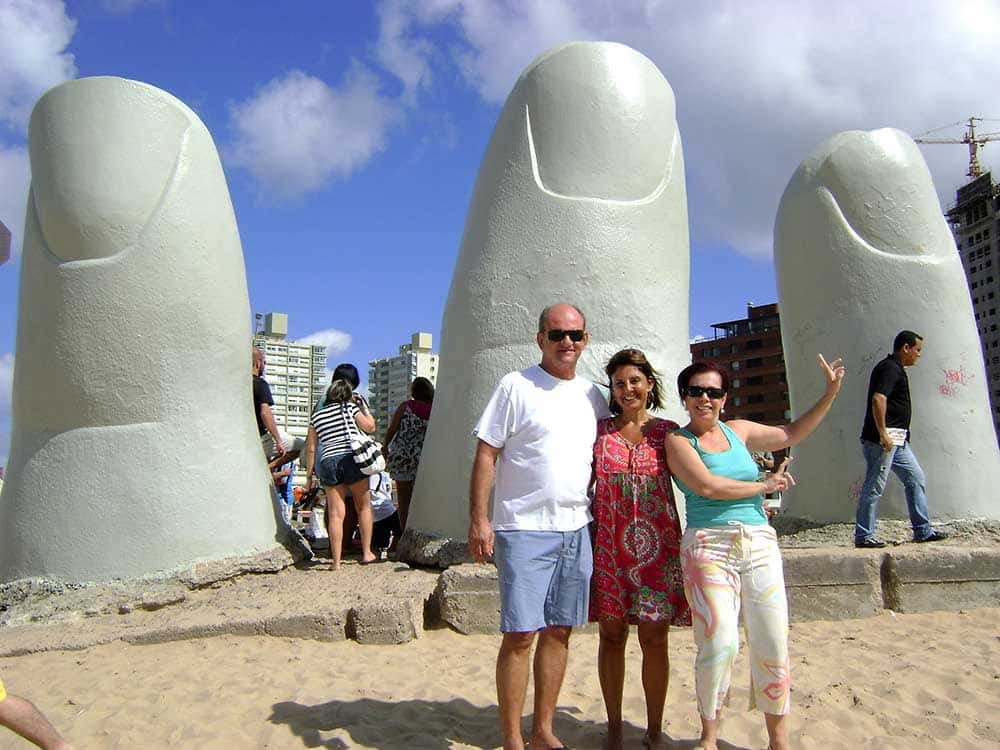 Duas mulheres e um homem em frente a dedos esculpidos e enterrados na areia em praia de Punta del Este