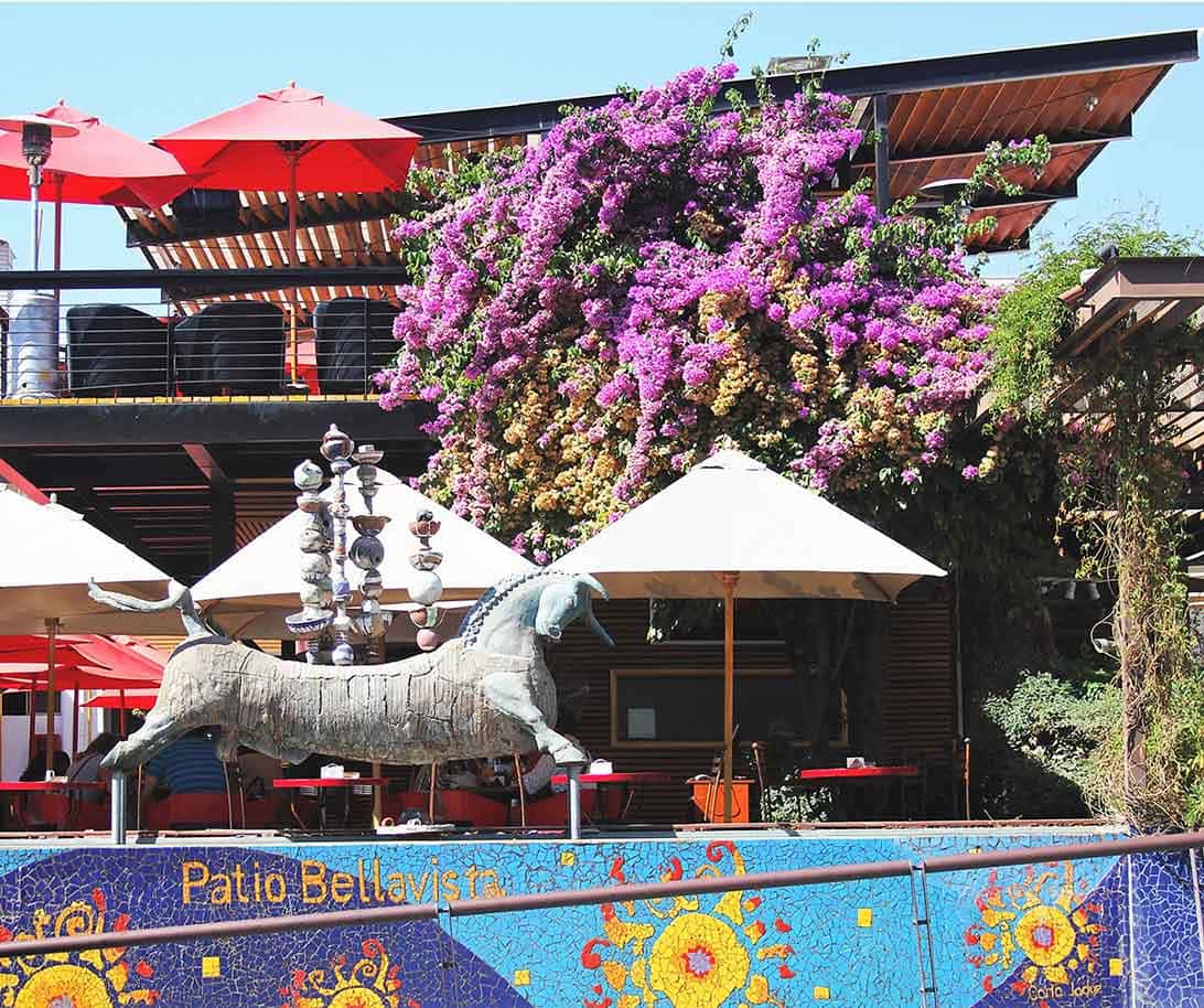 Muro colorido de azulejos com dizeres Patio Bellavista, estátua, mesas com sombrinhas, árvores com flores roxas: o que fazer em Santiago
