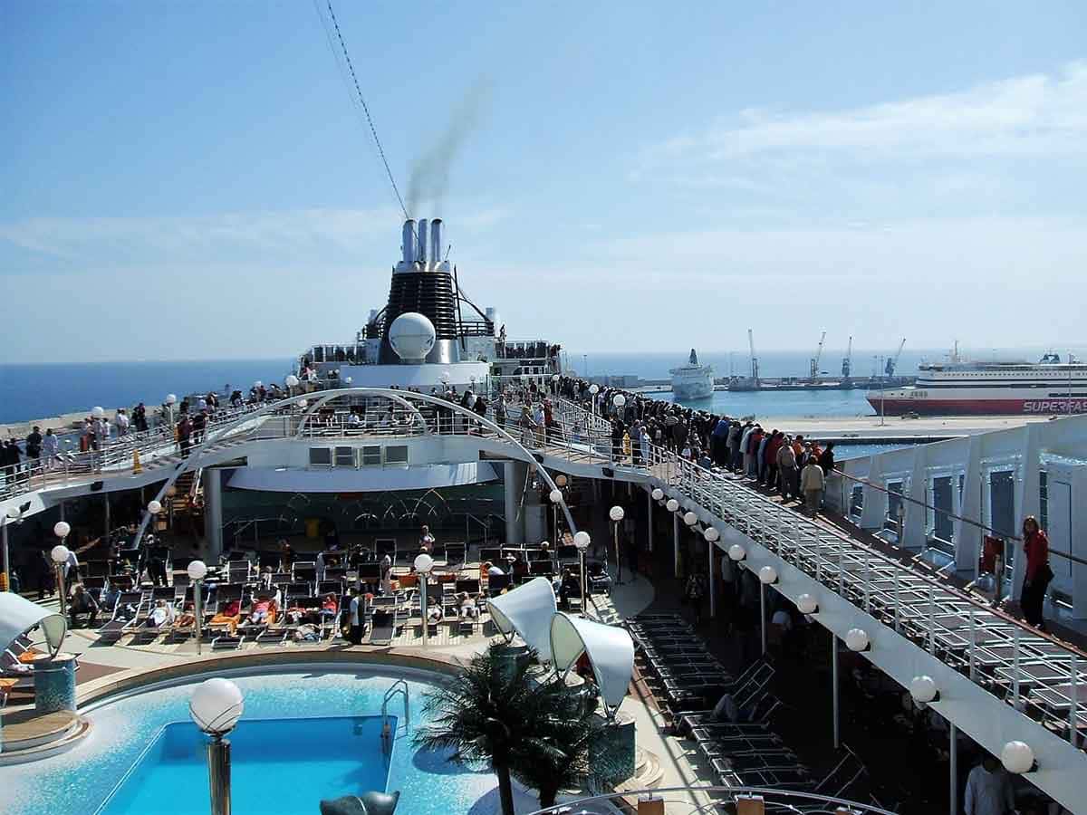 Viagem de navio: foto mostra interior do mesmo, com piscina e pessoas, a chaminé na proa e ao fundo, o porto