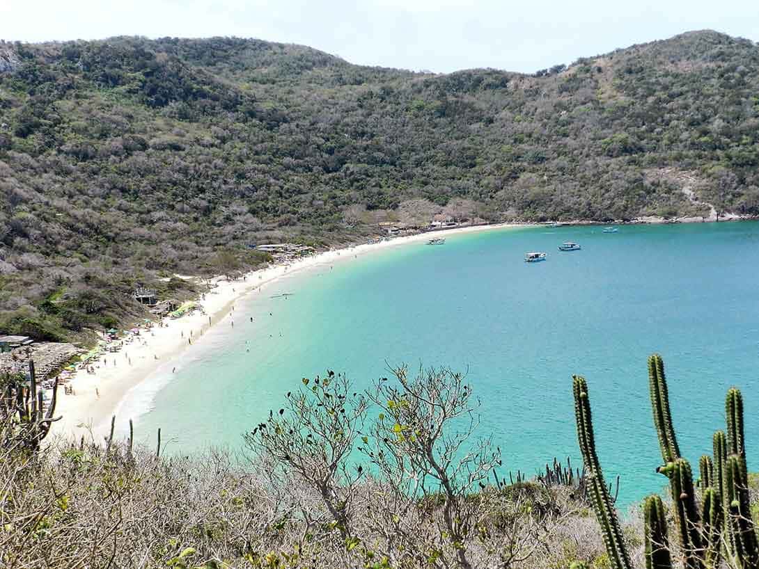 Praia com faixa de areia branca, água azul, barcos ao fundo, morro, chamada Praia do Forno e uma das praias de Arraial do Cabo