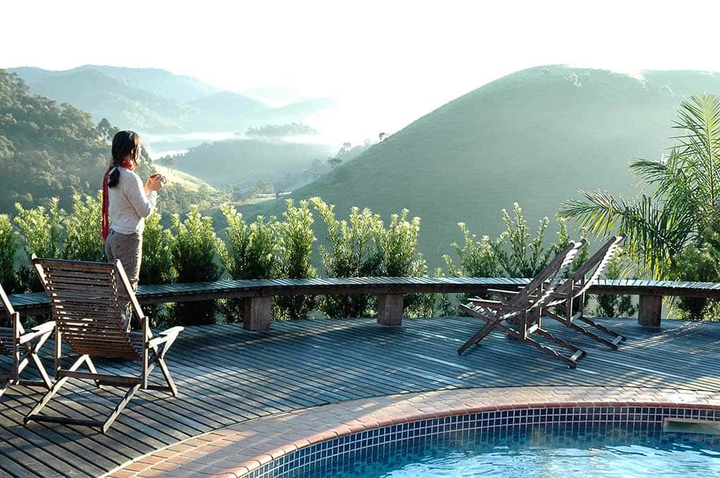 Moça tomando café, duas cadeiras e detalhe de uma piscina com montanhas ao fundo em uma das pousadas em Visconde de Mauá
