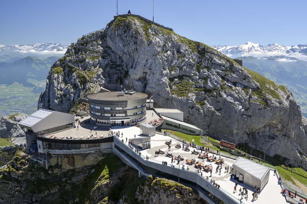Montanha com prédio redondo, plataforma de cimento onde se encontram várias pessoas, ferrovia com vagão ao fundo: Monte Pilatus em Lucerna, na Suíca