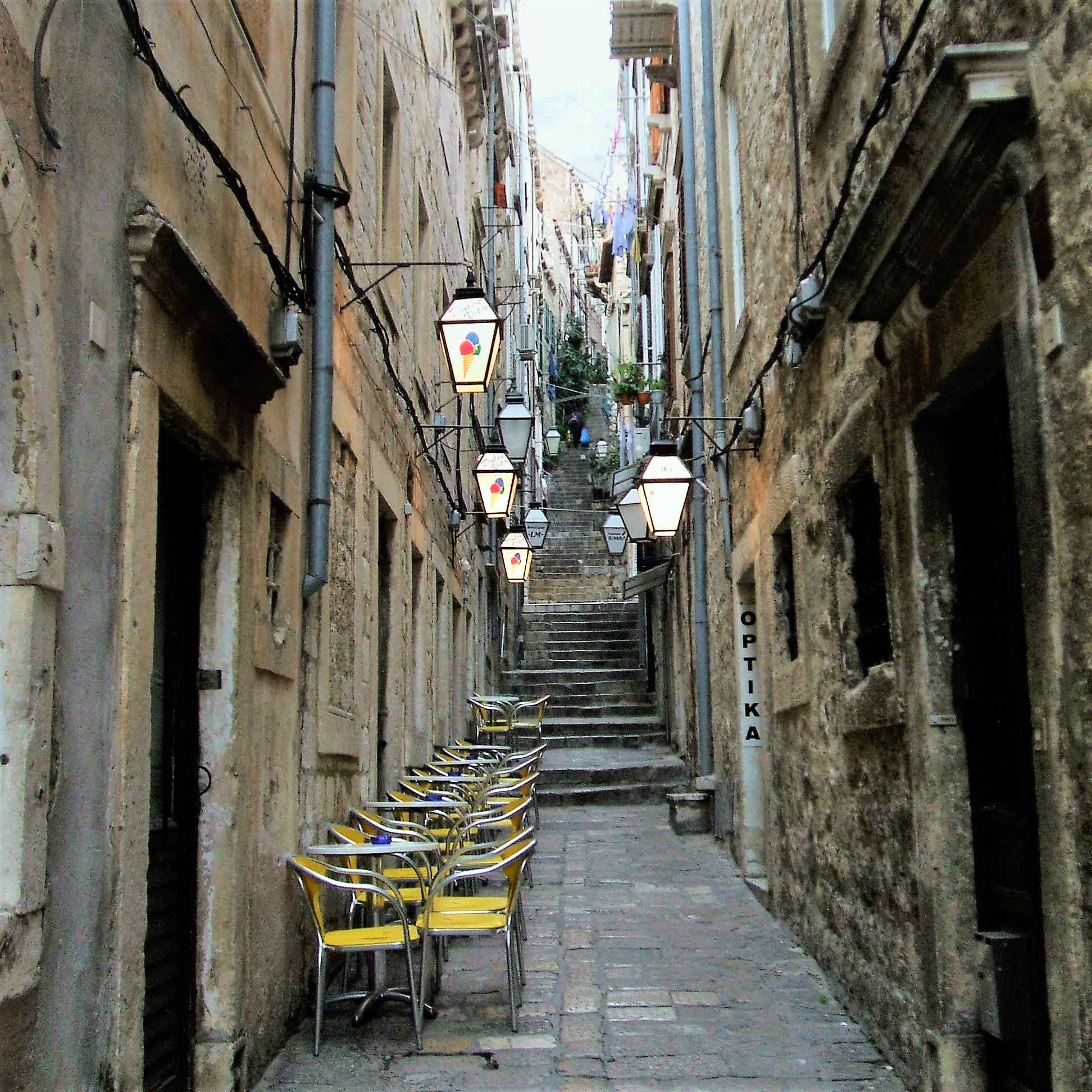 Em rua paralela, mesinhas à espera do freguês, lanternas e varais em Dubrovnik