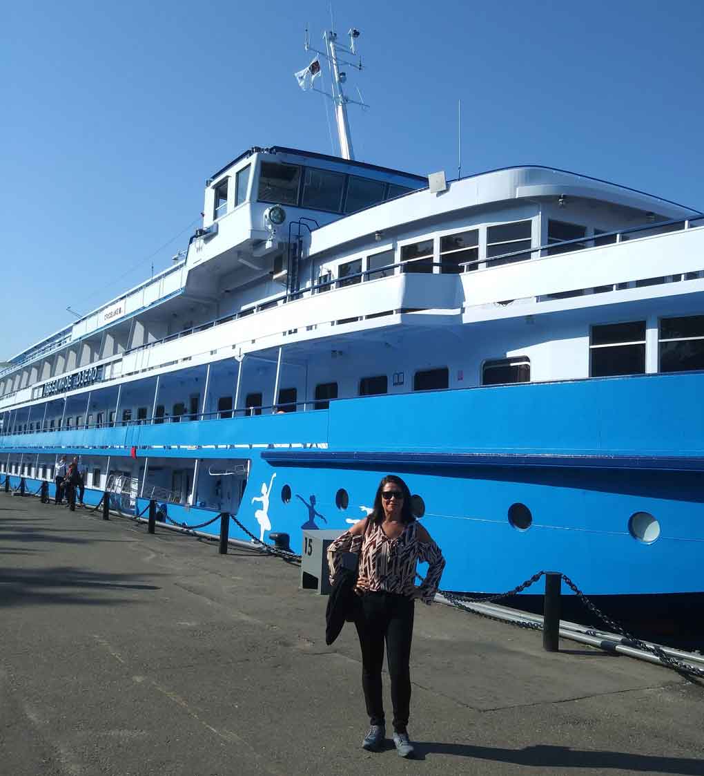 Moça posa em frente a barco azul e branco, que realiza cruzeiro pela Rússia
